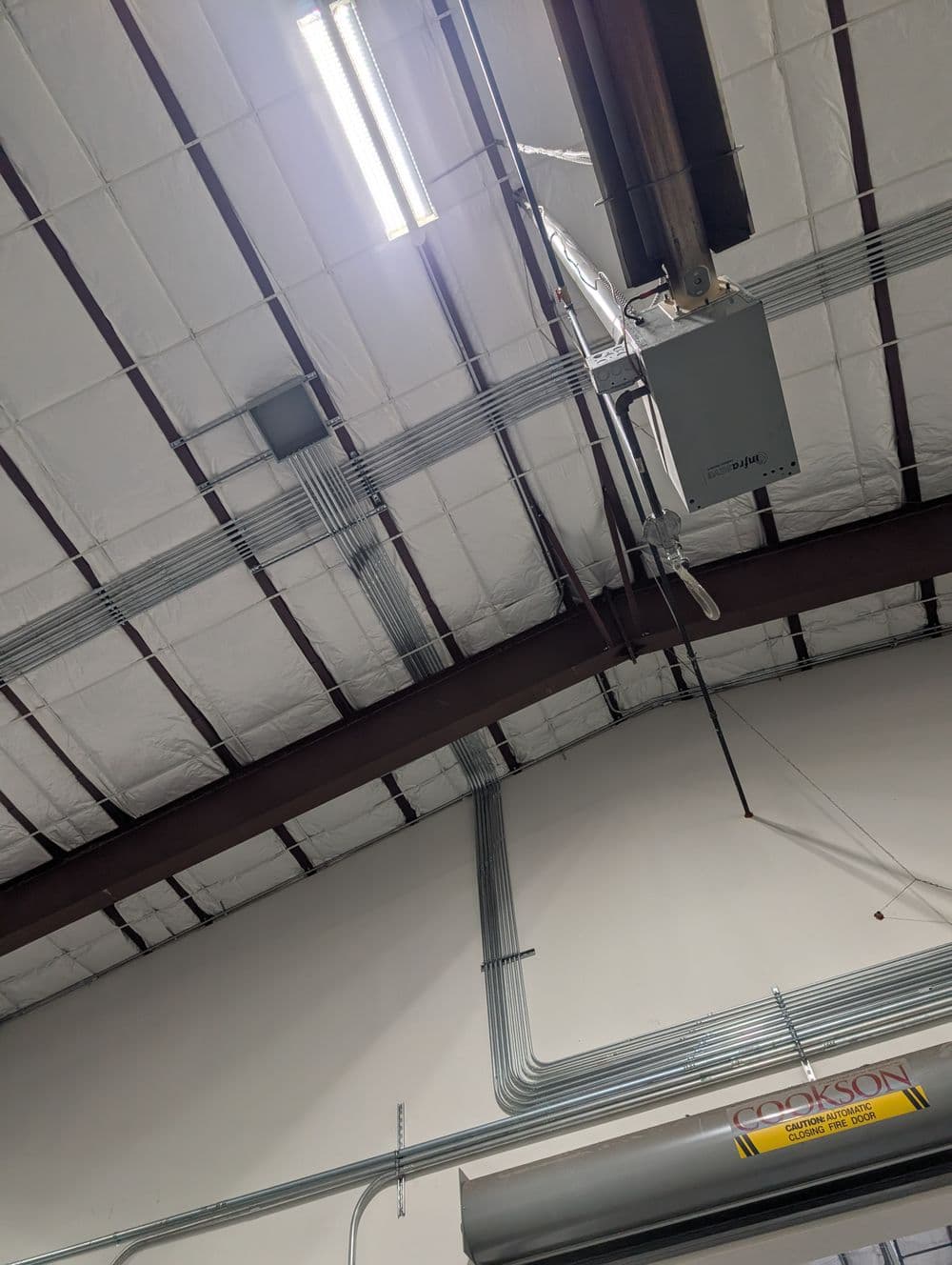 Warehouse ceiling with metal beams, lighting fixtures, and electrical conduits.